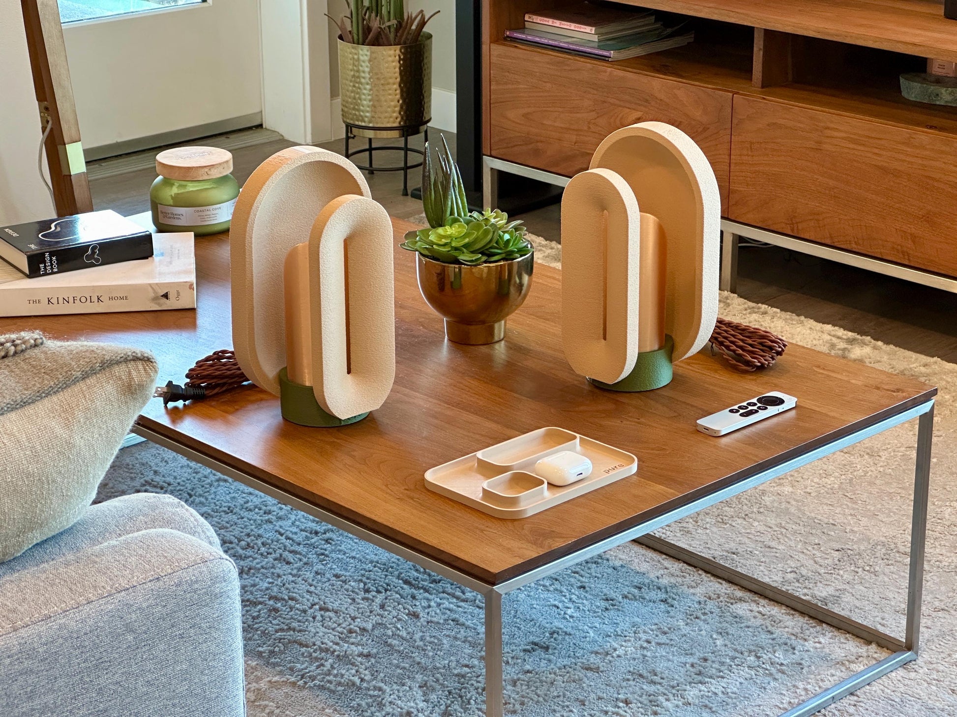 A modern dimmable table lamp with a stylish design, emitting a warm amber light, placed on a wooden surface surrounded by a plant, a reed diffuser, and a watch.