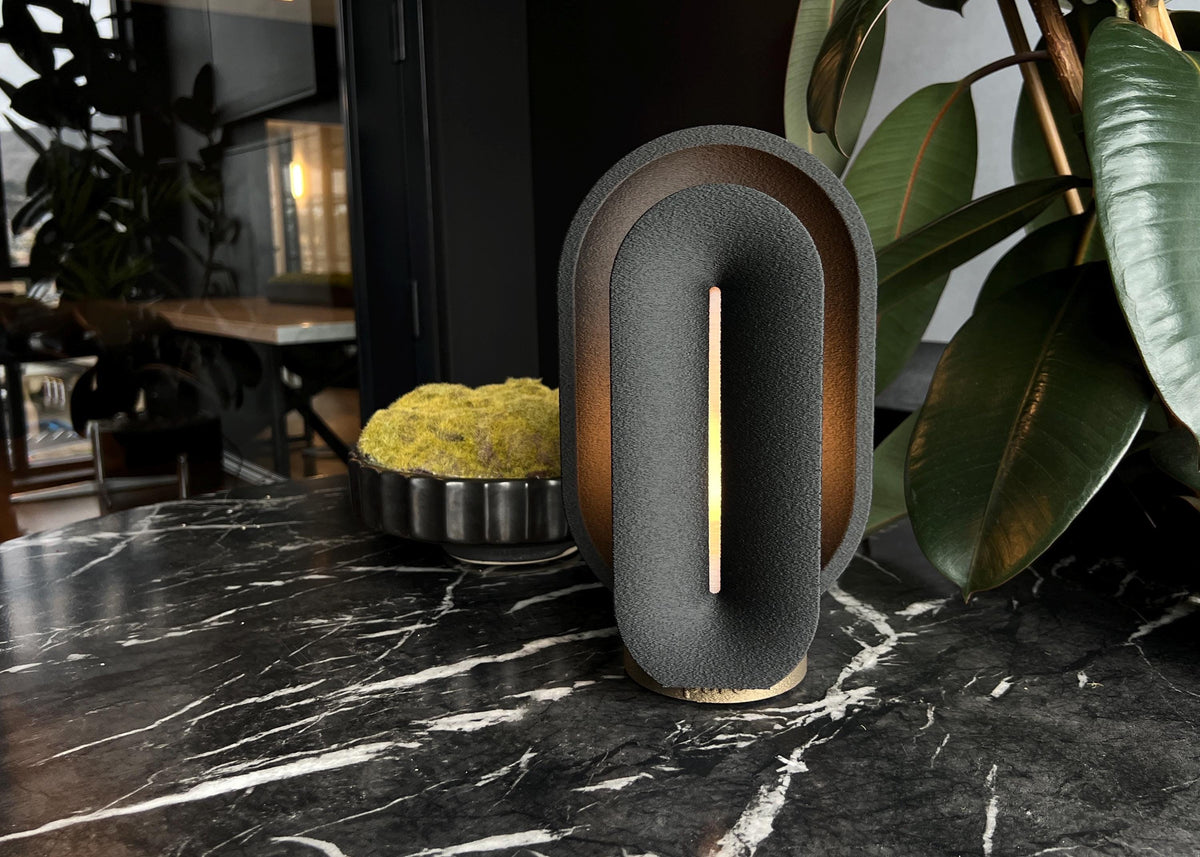 Black desk lamp glowing on a dark marble surface beside green plants, showing the sculptural Dune silhouette.