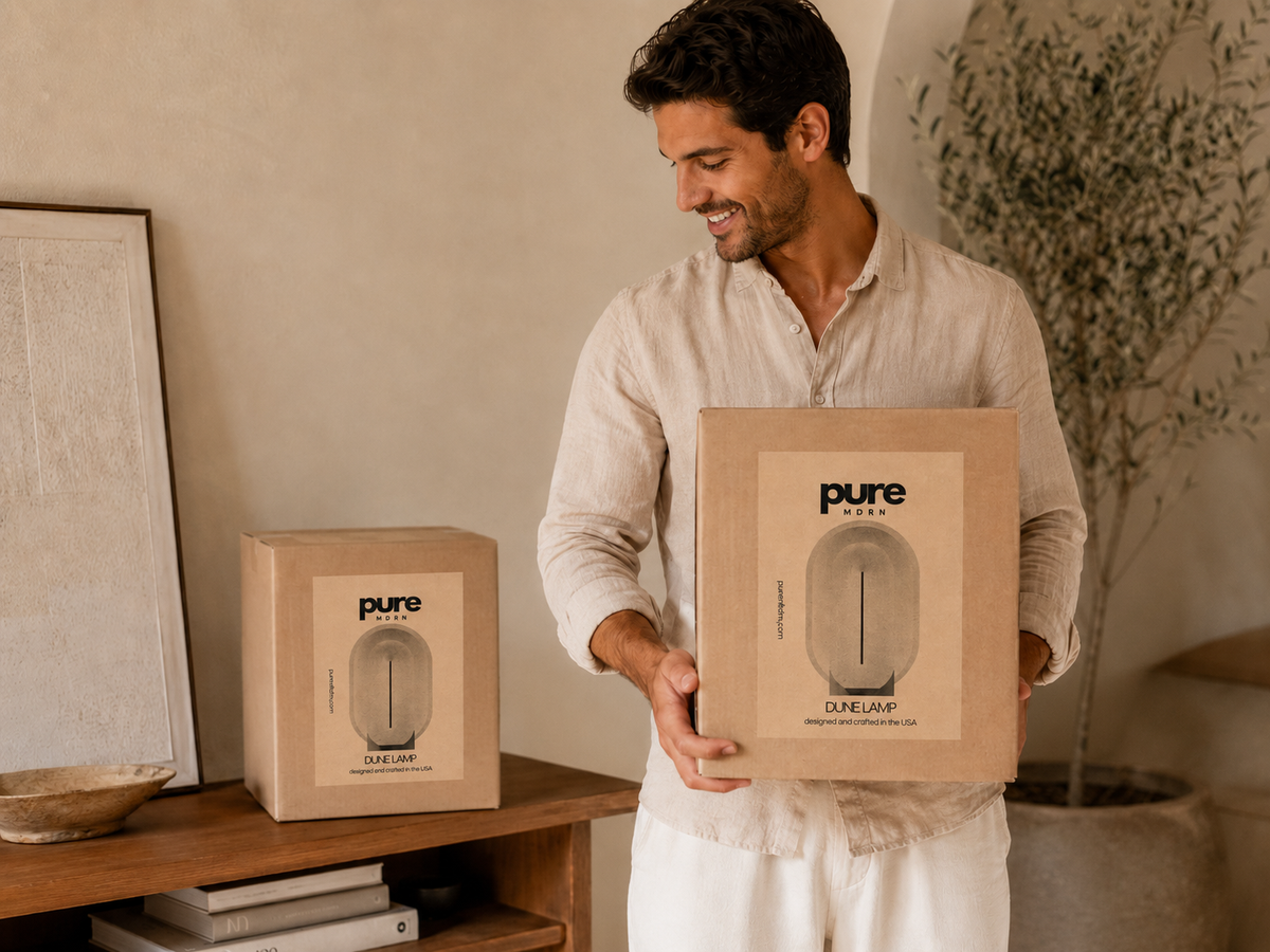 Man holding a PURE MDRN Dune table lamp shipping box, with a second branded package displayed on a wooden console.