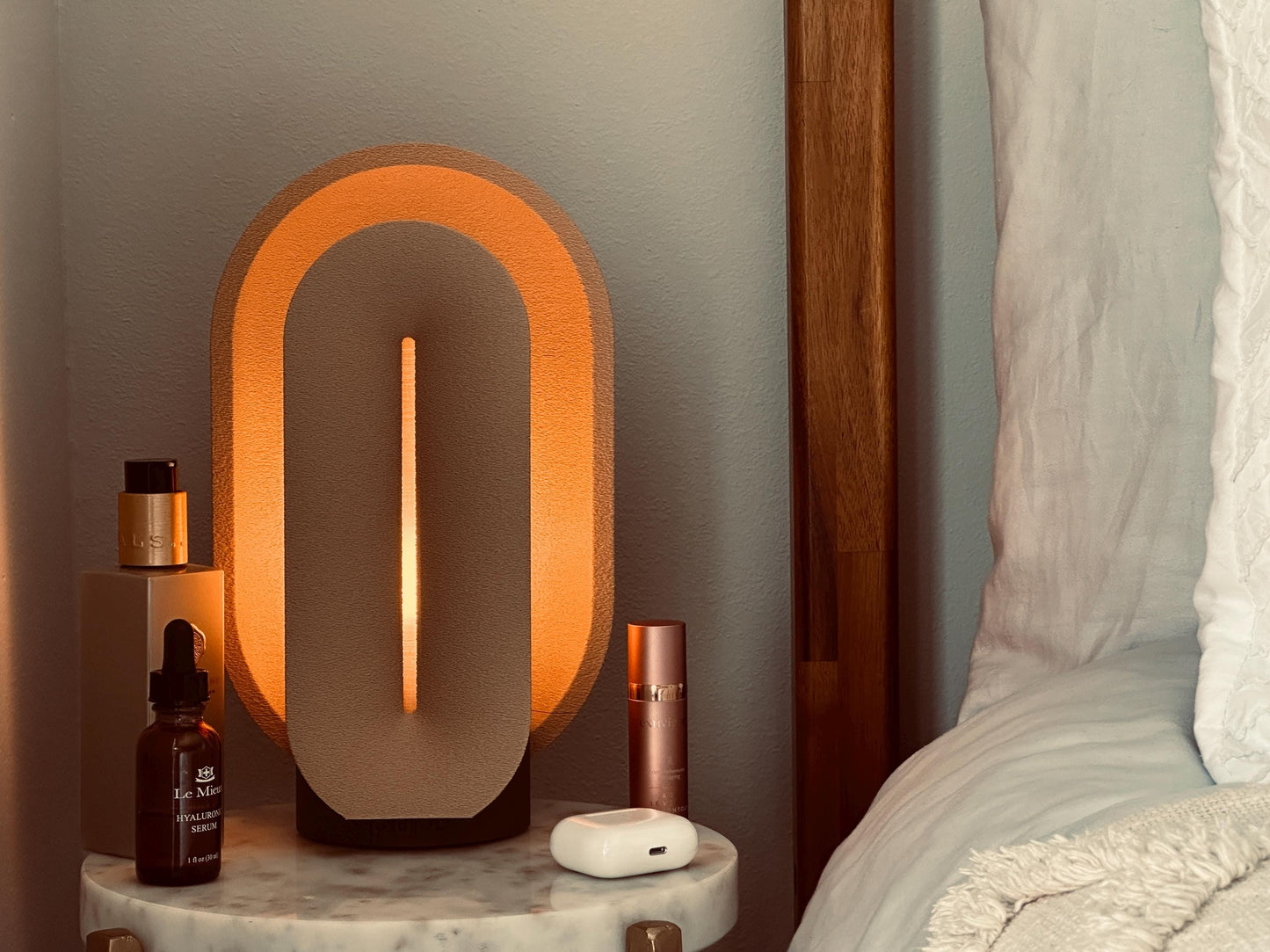 Beige bedroom lamp casting a warm glow from a compact bedside table beside neutral bedding and decorative objects.
