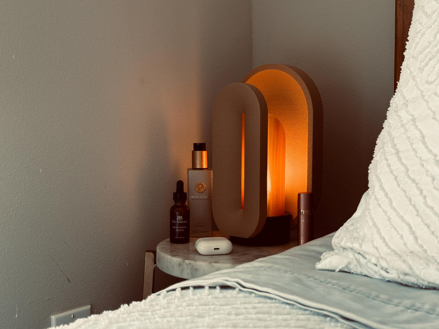 Sand small accent lamp creating warm mood lighting beside perfume bottles, a candle, and softly layered bedding.
