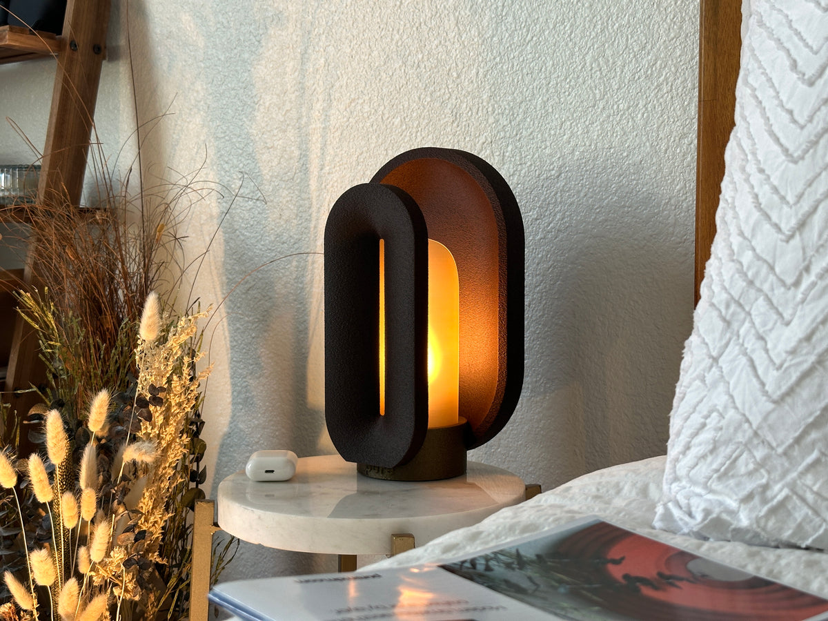 Coffee brown bedside lamp glowing beside white bedding, dried botanicals, and a magazine on a round marble nightstand.