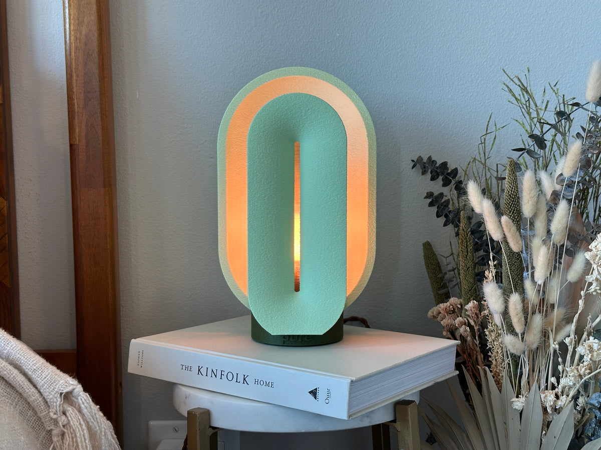 Matcha green small lamp glowing on stacked books beside dried flowers in a softly styled bedroom setting.